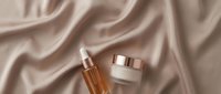 Luxury skincare products with golden serum dropper and cream jar displayed on draped beige silk fabric for cosmetics, beauty, and wellness marketing.