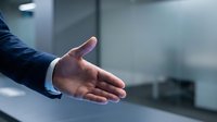 Businessman in dark suit extending open hand for handshake or greeting gesture against blurred office background, corporate partnership and business deal concept.