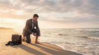 Professional businessman in suit sitting on beach at sunset with briefcase, contemplating work-life balance and career decisions by ocean waves.