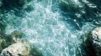 Aerial view of crystal clear turquoise ocean water surface with sunlight reflections creating mesmerizing patterns, rocky coastline visible along pristine tropical shore.