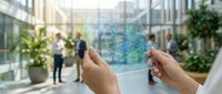 Hands holding transparent tablet displaying digital data analytics graphs in modern office lobby with business professionals in background.