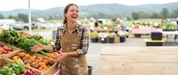 Young woman farmer in apron selling fresh organic vegetables at outdoor farmers market with mountain landscape background.