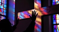 Silhouette of person with raised hand reaching toward wooden cross illuminated by colorful stained glass light in church interior for worship concepts.