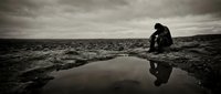 Person sitting alone by water puddle in barren landscape, reflecting on solitude and melancholy. Moody atmospheric scene for editorial, mental health awareness, or contemplative storytelling projects.