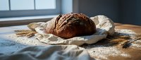 Freshly baked artisan dark bread loaf on linen cloth with wheat stalks and flour dust on wooden table in modern kitchen for bakery and cooking concepts.