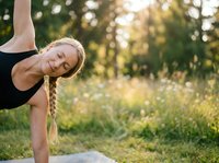 Young woman with blonde braided hair, Caucasian ethnicity, performing a yoga handstand pose outdoors in a sunny meadow with green trees and wildflowers in background.