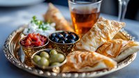 Mediterranean appetizer platter with phyllo pastries, assorted olives, roasted peppers, and dipping sauce served on decorative silver tray with glass of tea.