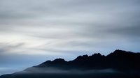 Silhouetted mountain range shrouded in dense fog and mist during dawn or dusk, creating a moody atmospheric landscape with layered peaks against a cloudy sky.