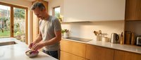 Middle-aged man with gray hair and light skin tone preparing food at kitchen counter with wooden cabinets and white countertop, natural light from garden doors.