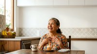 Senior woman with gray hair and warm smile enjoying breakfast at kitchen table with pastry and coffee, morning light streaming through window.