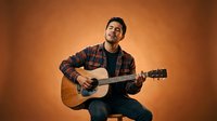 Young male musician with dark hair and facial hair, appearing to be of South Asian or Middle Eastern descent, playing acoustic guitar against warm orange studio background, wearing plaid shirt over black top.