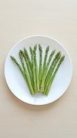 Fresh green asparagus spears arranged on white ceramic plate, overhead view on neutral beige background for healthy cooking and nutrition concepts.