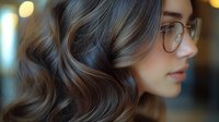 Profile view of young woman with wavy brown hair and glasses against blurred bokeh background, showcasing elegant hairstyle.