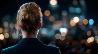 Woman in business attire viewed from behind facing blurred city lights and bokeh at night, creating atmospheric mood for leadership and future vision concepts.