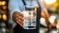 Glass of sparkling water with ice cubes held by hand against blurred warm bokeh background, refreshing drink served at restaurant or bar.