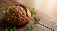 Freshly baked artisan sourdough bread loaf with crispy golden crust on rustic wooden table with fresh herbs for bakery and cooking concepts.
