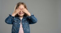Young blonde Caucasian girl covering eyes with hands wearing denim jacket and striped shirt on gray background for childhood emotions and playful concepts.