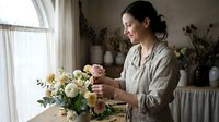 Young woman florist arranging fresh flower bouquet in bright studio workspace. Professional flower arrangement and botanical design concept.