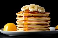Stack of fluffy pancakes topped with fresh banana slices and dripping maple syrup on white plate against dark background, with citrus fruits beside.