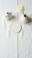 Minimalist ceramic vases and empty plate arranged on white linen fabric with fresh cream ranunculus flower, styled for home decor and interior design projects.