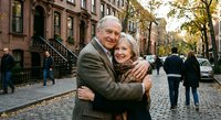 Happy senior Caucasian couple embracing on autumn city street with historic brownstone buildings. Elderly man and woman showing affection outdoors.