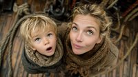 Young Caucasian mother and toddler son wearing cozy knit scarves looking up with surprised expressions, family winter portrait from above on wooden background.