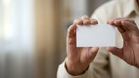 Close-up of male hands holding blank white business card against blurred neutral background, suitable for corporate branding and professional networking materials.