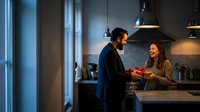 Young couple enjoying morning coffee together in modern kitchen with pendant lighting and marble countertops creating intimate domestic atmosphere.