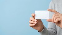 Close-up of hands holding blank white business card against light blue background, suitable for branding, corporate identity, and marketing mockup presentations.