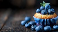 Fresh blueberry muffin topped with ripe berries and mint leaf on rustic wooden background, selective focus creates appetizing dessert composition.