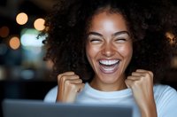 Young African American woman with natural curly hair laughing joyfully while looking at laptop screen, expressing genuine happiness and excitement in evening setting.