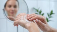 Young woman with fair skin applying moisturizing cream to her hands in front of a mirror, demonstrating skincare routine and hand care product application.
