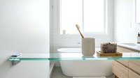 Minimalist bathroom shelf with ceramic toothbrush holder, potted plant, and wooden accessories on glass shelf above white sink.