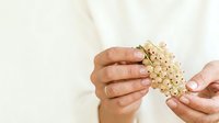 Young adult with medium skin tone holding handful of small beige pearls or decorative beads against neutral background, suitable for jewelry, beauty, luxury, and design projects.