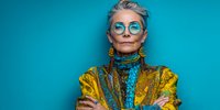 Senior woman with short platinum hair wearing blue round glasses, colorful yellow and turquoise outfit, and statement earrings against vibrant blue background.