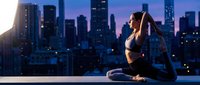 Young athletic woman practicing yoga meditation pose on urban rooftop terrace at sunset with city skyline background for wellness and fitness lifestyle.