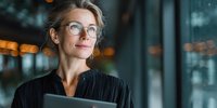 Confident mature businesswoman with glasses holding digital tablet in modern office environment with blurred city lights background.