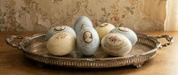 Vintage decorated Easter eggs displayed in ornate metal tray on wooden surface with floral wallpaper background for spring holiday celebrations.