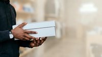 Man with dark skin tone wearing formal business attire holding white envelope or document box in office setting, suitable for corporate communications and business concepts.