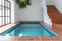 Indoor swimming pool with turquoise water in minimalist white room featuring wooden staircase and large windows with natural light.