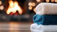Cozy stacked bath towels in white and teal colors against blurred fireplace background with warm bokeh lights creating relaxing spa atmosphere.