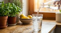 Fresh water being poured from glass bottle into drinking glass on wooden kitchen counter with green herbs and fruits in warm sunlight.