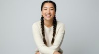 Young Asian woman with braided hair wearing cream knit sweater, smiling at camera against white background. Portrait for lifestyle, fashion, and wellness content.