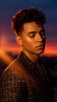 Young mixed-race man with curly hair in sparkly jacket during golden hour sunset, eyes closed in peaceful expression against dramatic colorful sky backdrop.