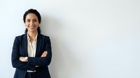 Professional woman with dark hair and olive complexion wearing navy blazer and white shirt, arms crossed, standing against white background for corporate portraits and business headshots.