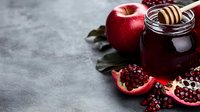 Fresh red apple, dark honey jar with wooden dipper, and cut pomegranate on gray concrete background with copy space. Traditional Rosh Hashanah symbols.