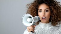Young woman with curly brown hair and olive complexion holding white megaphone near face, expressing excitement and announcement. Suitable for marketing, promotion, communication campaigns.