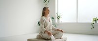 Young Caucasian woman in white clothing practicing meditation in bright minimalist room with plants by window for wellness and mindfulness lifestyle.