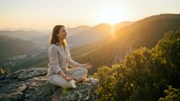 Young woman meditating on mountain cliff at sunrise with golden light and scenic valley view. Peaceful mindfulness practice in nature for wellness.