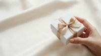 A person's hand holding a small white gift box with a gold ribbon bow against a soft, neutral background with natural lighting.